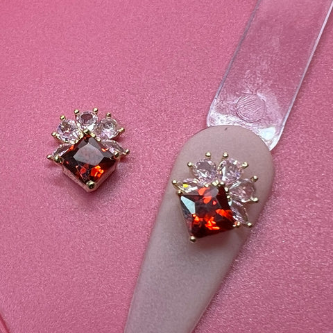 A pair of ornate earrings featuring square red gemstones encircled by clear marquise-cut stones are displayed on a pink surface. One earring lies directly on the pink background, while the other, reminiscent of Dynamic Nail Supply’s 2 pcs Luxury 3D Charm for Nails Art Design with Pink and Red Rhinestone Engraved, is perched on a translucent clip similar to nail art design. The stones are securely held in place by gold prongs.
