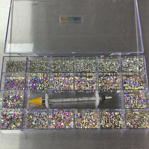 The Dynamic Nail Supply [AB Iridescent gold-back] Rhinestones set includes a clear plastic storage box with 20 compartments filled with various shapes like stars, hearts, and circles. It comes with a rhinestone applicator pen featuring a yellow tip, and the lid prominently displays the word Dumonic.