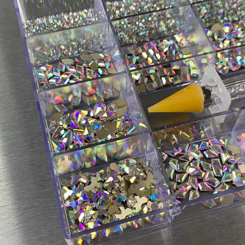 A close-up of a Dynamic Nail Supply compartmentalized tray showcases various AB iridescent gold-back rhinestones featuring big shapes like hearts and stars. Next to the tray, an orange applicator sits on a metallic surface, reflecting light to create colorful patterns, from the [AB Iridescent gold-back] Rhinestones set collection with 20 cells including 14 big shapes and 6 round shapes.