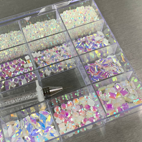 The [White Pearl Iridescent] Rhinestones set by Dynamic Nail Supply includes a plastic organizer box containing a variety of colorful rhinestones in white pearl iridescent. This set features shapes such as stars, squares, and hexagons, along with an applicator pen positioned gracefully on the left side of the box against a metallic surface.