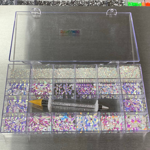 A clear plastic organizer box from Dynamic Nail Supply displays the [White Pearl Iridescent] Rhinestones set, which includes 14 big shapes and 6 round shapes of nail art gems. A dual-ended picker tool with a yellow tip is nestled inside. The lid is slightly open, exposing a metallic surface background and segments of additional boxes.