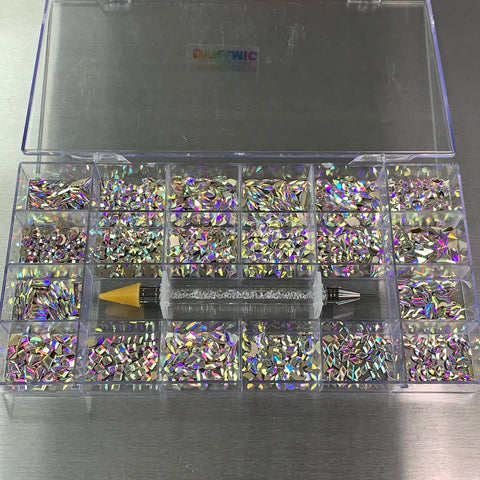 A clear plastic box from Dynamic Nail Supply's AB Iridescent gold-back Rhinestones set showcases multiple compartments filled with shimmering rhinestones in 20 big shapes. A rhinestone picker tool with a yellow tip lies across the box, which sits on a metallic surface. The lid displays a subtle, colorful logo.