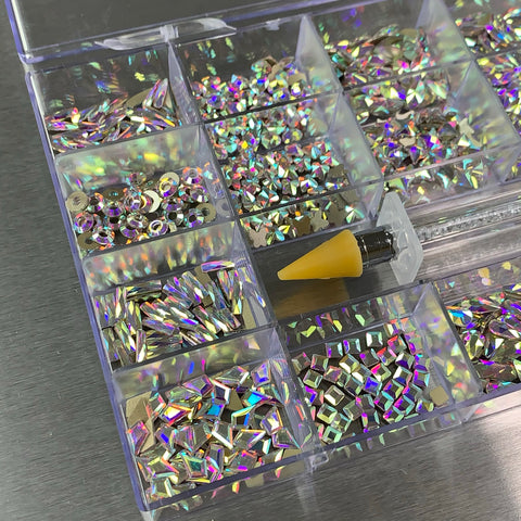 The [AB Iridescent gold-back] Rhinestones set from Dynamic Nail Supply features a clear plastic organizer containing 20 different big shapes, with 50 pieces each. Excluded from this collection are six round shapes. This essential nail supply includes iridescent rhinestones stored in nine compartments, showcasing a variety of sizes and designs. A small tool with an orange grip is conveniently placed on one of the compartments, while the metallic, reflective surface beneath enhances its elegant appeal.