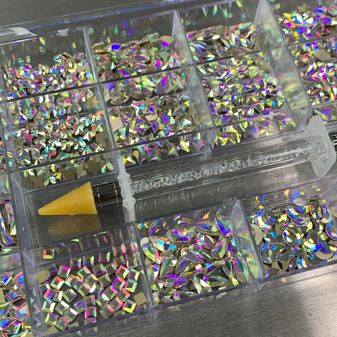 A chic clear plastic organizer from Dynamic Nail Supply, ideal for any nail supply collection, neatly holds the [AB Iridescent gold-back] rhinestones set in various shapes like rectangles and teardrops. A rhinestone picker with a yellow tip and a clear handle is positioned diagonally across the box, while the reflective surface beneath enhances their brilliance.