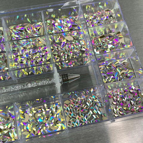 Introducing the [AB Iridescent gold-back] Rhinestones set by Dynamic Nail Supply: A transparent box containing a variety of rhinestones in 20 unique large shapes, with 50 pieces of each shape. Excluding six round shapes, these stones showcase highly reflective surfaces that emit multicolored patterns, making them ideal for any nail supply collection. Also included is an elegant rhinestone picker tool featuring a crystal handle, placed conveniently beneath the box.