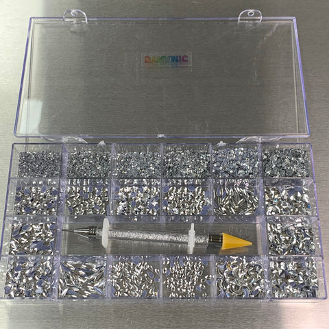 A clear plastic organizer box with an open lid showcases 14 large shapes and 6 round shapes of silver silver-back rhinestones in separate compartments. A rhinestone applicator tool with a white barrel and a yellow tip lies across the middle, while Dynamic Nail Supply is printed on the lid in rainbow colors.