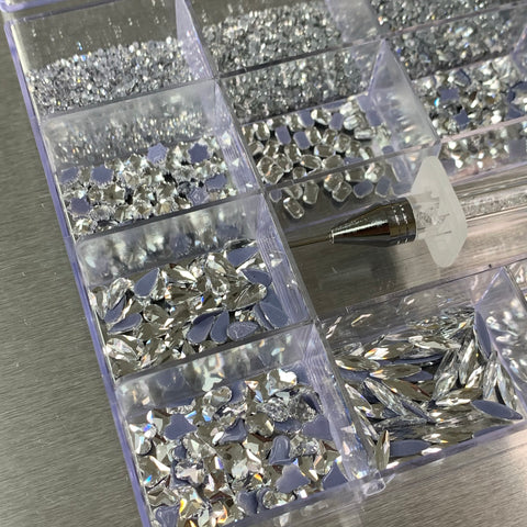 A [Silver silver-back] Rhinestones set from Dynamic Nail Supply includes a 20-compartment clear organizer box filled with assorted shiny silver rhinestones in various shapes, such as big round, teardrop, and marquise. A convenient rhinestone picker tool rests on the edge against a metallic background.