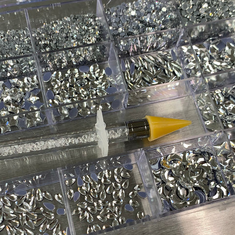 A yellow-tipped rhinestone applicator pen rests on a tray filled with Dynamic Nail Supply's [Silver silver-back] Rhinestones set, featuring various silver pieces in 14 big shapes and 6 round shapes, all neatly organized in a 20-cell box. The pen's clear barrel houses tiny rhinestones, creating an inviting craft or jewelry-making setup.