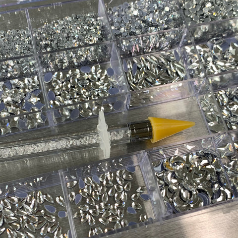 The Dynamic Nail Supply [Silver silver-back] Rhinestones set comes in a 20-compartment clear box, filled with assorted silver rhinestones in various shapes and sizes, including 14 big shapes. At the center of the set is a rhinestone pick-up tool, featuring a cylindrical body filled with small crystals, a clear handle, and topped with a yellow tapered cap on one end.