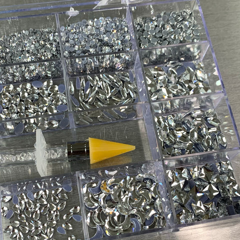 The [Silver silver-back] Rhinestones set from Dynamic Nail Supply includes a 20-compartment plastic organizer containing 14 big shapes and 6 round shapes of silver rhinestones. The box is topped with a tool featuring a yellow tip, ideal for picking up and applying the glittering gems on a metallic background.