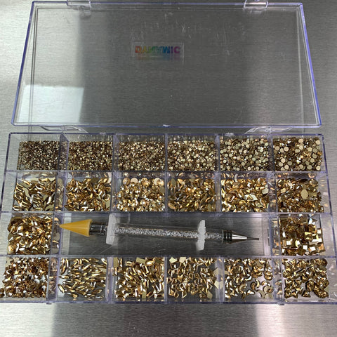 A clear plastic organizer box from Dynamic Nail Supply, labeled "Champagne Gold" Rhinestones Set - 20 Cells Box, contains various nail art embellishments, including 14 big shapes and 6 round shapes, sorted into separate compartments. A rhinestone-studded pen with a yellow tip is placed across the sections. The lid displays a blurred Dynamic Nail Supply logo with colorful text on a metallic surface background.