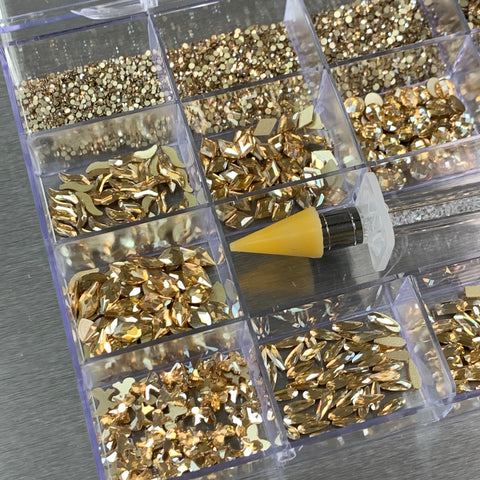 The [Champagne Gold] Rhinestones set by Dynamic Nail Supply is displayed in a transparent organizer featuring 20 compartments filled with dazzling nail art decorations, including 14 large shapes and 6 round shapes. In the center of the arrangement, a small tube of plastic nail glue with a yellow cap sits atop one of the compartments—an ideal addition to any nail supply collection.