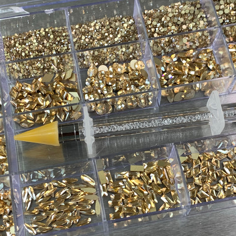 A plastic container from Dynamic Nail Supply, known as the [Champagne Gold] Rhinestones set, features 20 cells containing an assortment of rhinestones in 14 big shapes and 6 round shapes. Resting on top is a pen-like tool, crucial for any nail supply kit, distinguished by its yellow pointed tip and a clear section filled with small crystals. The backdrop is a reflective metallic surface.