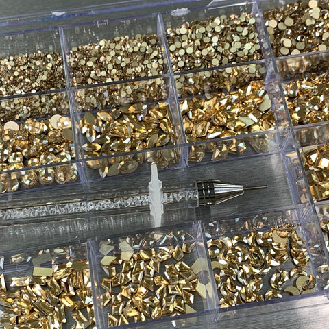 The [Champagne Gold] Rhinestones set by Dynamic Nail Supply features a clear plastic storage box with 20 compartments filled with diverse sizes and shapes of sparkling, champagne gold rhinestones. This essential nail supply also includes a rhinestone picker tool with a translucent handle and a metal tip, conveniently positioned diagonally across the compartments.