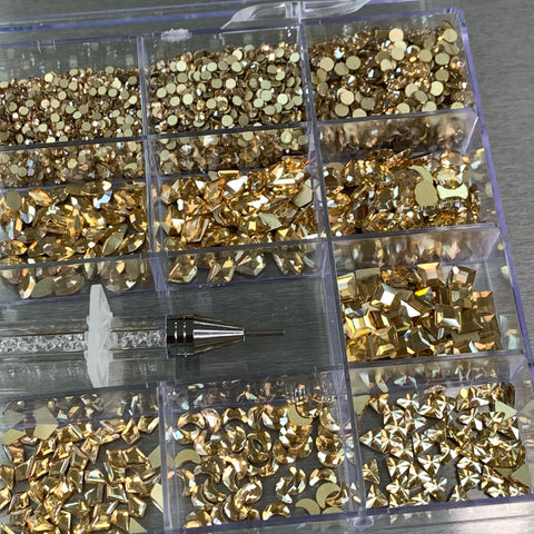 A [Champagne Gold] Rhinestones set from Dynamic Nail Supply features a 20-cell box with a stunning nail supply of rhinestones, beautifully sorted into compartments. These include 14 big shapes such as stars and squares, along with 6 round shapes. Paired with the set is a clear-handled tool for applying the rhinestones, providing everything you need for creating dazzling nails.