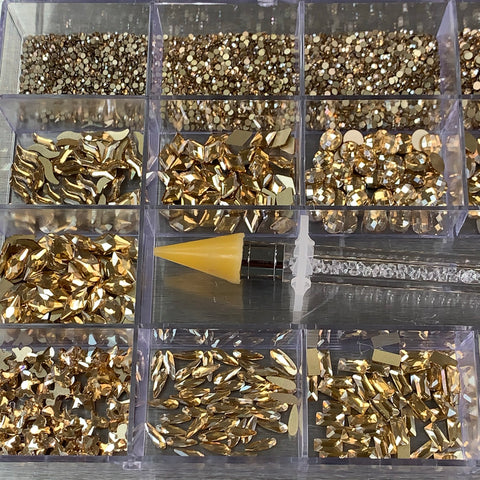 A 20-cell clear plastic organizer from Dynamic Nail Supply, containing the [Champagne Gold] Rhinestones set with 14 big shapes and 6 round shapes, is filled with shimmering champagne gold rhinestones. Positioned diagonally across the middle of the organizer on a metallic surface is an essential nail supply tool—a rhinestone picker featuring a yellow tip.