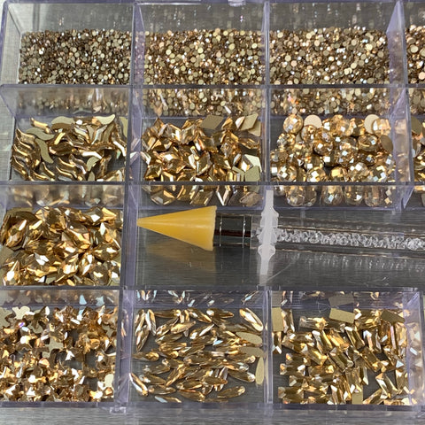 A plastic organizer featuring a set of [Champagne Gold] rhinestones from Dynamic Nail Supply boasts an assortment of 14 big shapes and 6 round shapes, all housed in a 20-cell box. Lying across the center compartments is a rhinestone picker tool with a yellow, cone-shaped tip and shiny metallic finish, ideal for nail supply enthusiasts. The reflective surface backdrop enhances this elegant display.