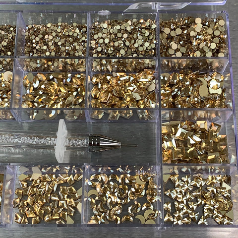 A plastic organizer box from Dynamic Nail Supply holds an assortment of Champagne Gold nail art embellishments, featuring 14 big shapes such as circles, squares, and diamonds. The compartments are arranged in a grid layout. A nail art tool with a white handle and metal tip is placed across several sections of this stunning [Champagne Gold] Rhinestones set.