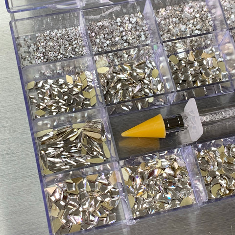 A Dynamic Nail Supply plastic organizer comes with the [Platinum gold-back] Rhinestones set, featuring 20 compartments filled with gleaming silver rhinestones and embellishments in 14 big shapes and 6 round shapes. A rhinestone picker with a yellow tip is positioned on top of the container, resting on a metallic surface.