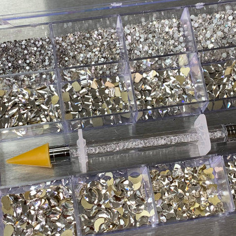 A [Platinum gold-back] Rhinestones set from Dynamic Nail Supply, featuring a combination of 14 big shapes and 6 round shapes, is neatly arranged in a transparent 20-cell box on a metallic surface. Accompanying the set is a crystal pen with a yellow cap, resting diagonally on the box and ideal for crafting or enhancing your nail supply essentials with added sparkle.