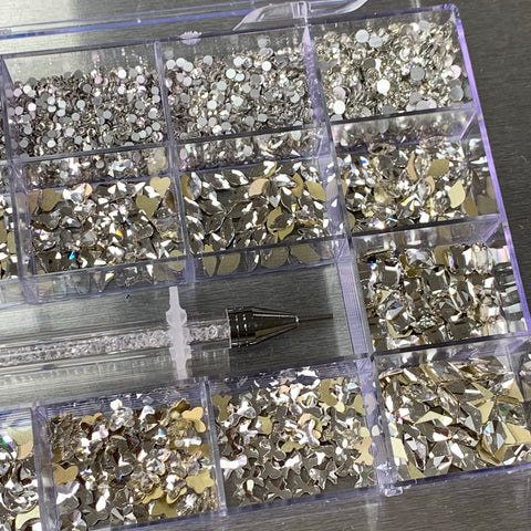 The [Platinum gold-back] Rhinestones set by Dynamic Nail Supply comes in a 20-celled plastic organizer box, showcasing a collection of big and round shapes in various forms. The compartments exhibit circular, teardrop, and irregular rhinestones with platinum gold-backs. Nearby is a nail supply tool featuring a clear handle and metal tip.