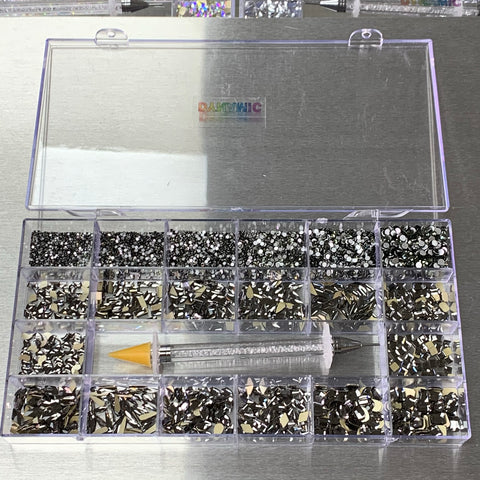 The [Titanium gold-back] Rhinestones set from Dynamic Nail Supply comes in a clear plastic organizer box with 20 compartments, featuring an impressive collection of rhinestones in 14 big shapes and 6 round shapes. Available in black, gray, and silver hues, it's perfect for all your nail supply needs. It also includes a rhinestone picker tool with a yellow tip. The background showcases a striking metallic surface.
