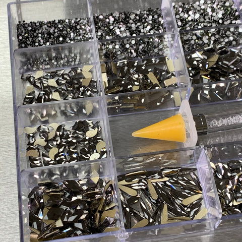 A close-up of the [Titanium gold-back] Rhinestones set by Dynamic Nail Supply showcases a 20-cell organizer box filled with black and silver rhinestones in various shapes, including 14 big styles and 6 round designs. Among these are round, teardrop, and angular forms. A yellow-tipped applicator is positioned at the bottom right of the image.