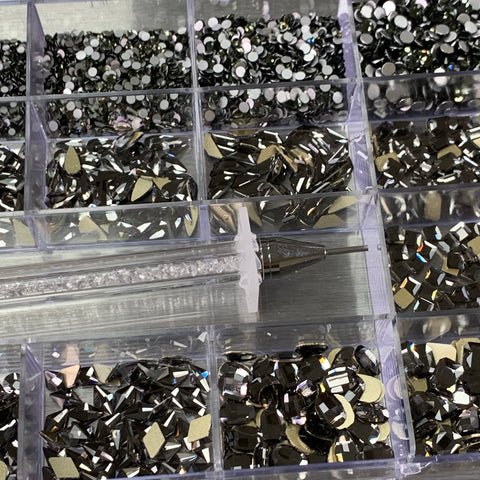 This close-up features the [Titanium gold-back] Rhinestones set by Dynamic Nail Supply, displayed in a 20-cell box containing an assortment of 14 big shapes and 6 round shapes in black and silver. A must-have for nail enthusiasts, the rhinestone picker tool with a clear handle rests on the edge, highlighting their shimmering surfaces.