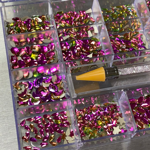 A compartmentalized storage box from Dynamic Nail Supply features a set of colorful and shiny [Amethyst] Rhinestones in various shapes, such as crescent moons and stars. The sections boast an array of bright pink, gold, and iridescent pieces. Additionally, a yellow-tipped tool designed for picking up the decorations is included in the box.