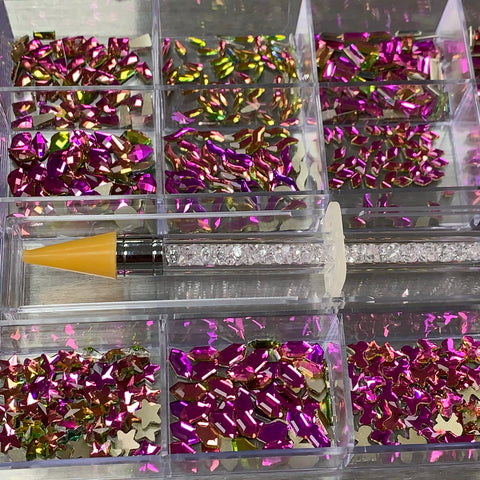 A divided container features the vibrant assortment of [Amethyst] Rhinestones from Dynamic Nail Supply, including stunning shapes like stars and teardrops. A rhinestone-studded picker tool with a yellow tip elegantly extends across the center, reflecting the brand's sophistication on a gleaming surface.