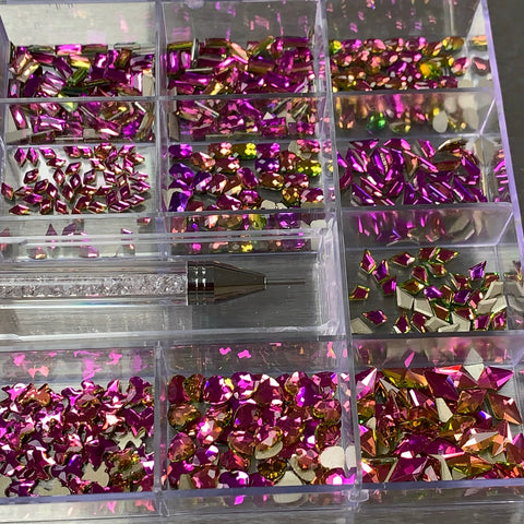 The [Amethyst] Rhinestones set by Dynamic Nail Supply includes a clear organizer with nine sections, each filled with iridescent pink and amethyst rhinestones. A sparkling pen with a transparent barrel is elegantly placed across the center. The rhinestones reflect pink, yellow, and green hues, resulting in a vibrant and colorful display.