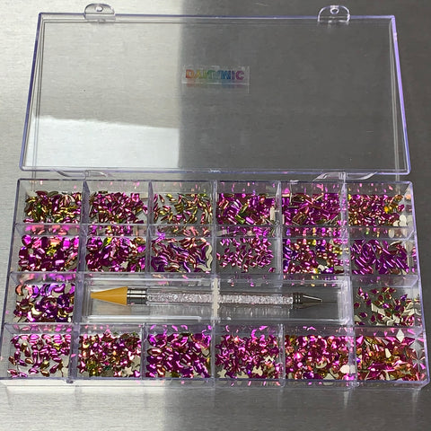 A clear organizer box with 16 compartments contains pink and gold iridescent nail art pieces. A nail art pen adorned with amethyst rhinestones and a yellow grip is positioned diagonally across the center on a metallic surface. The lid showcases a vibrant label from Dynamic Nail Supply, ideal for enhancing any nail supply collection.