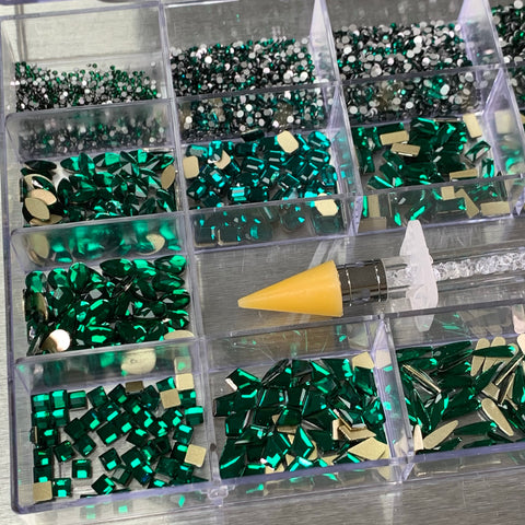 A divided plastic container from Dynamic Nail Supply contains a selection of Emerald Green rhinestones in various big and round shapes, offered in both green and silver. The set includes 14 big shape varieties like squares and rectangles, as well as 6 round shapes. A rhinestone applicator with a yellow tip is neatly placed on the edge of the container, adding convenience to this gleaming assortment.
