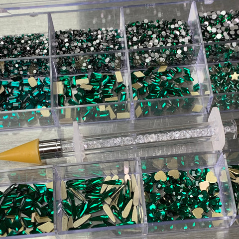 An Emerald Green Rhinestones set from Dynamic Nail Supply, featuring a 20-cell box with 14 big shapes and 6 round shapes, contains rhinestones in emerald green and silver colors in various designs, including round, heart, and rectangular. A rhinestone picker tool with a yellow tip is placed across the box, showcasing its clear handle filled with small, transparent rhinestones.