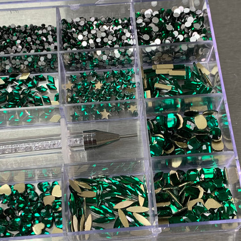 An organizer made of plastic containing compartments that are filled with [Emerald Green] rhinestones in a variety of shapes, including 14 big ones and 6 round ones. A transparent-handled rhinestone picker tool from Dynamic Nail Supply is laid across the compartments on a metallic surface.