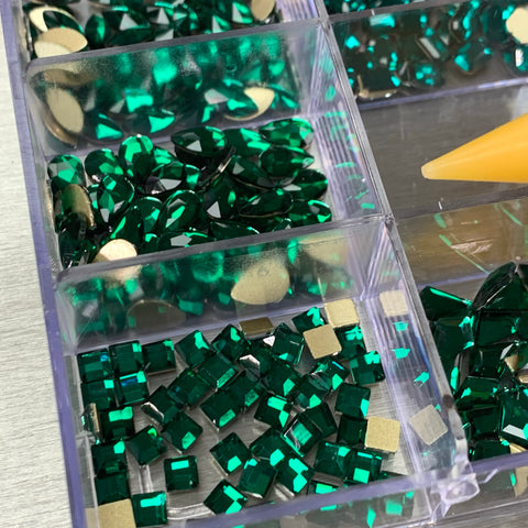 A plastic organizer with 20 compartments contains a variety of emerald green rhinestones from Dynamic Nail Supply, featuring 14 large shapes and 6 round shapes, all with metallic gold backing. A hint of a yellow pen tip is visible on the right side against a light-colored background. This set is ideal for enhancing your Dynamic Nail Supply collection with some extra sparkle.