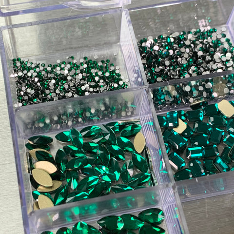 A close-up image of the Dynamic Nail Supply Emerald Green Rhinestones set, featuring various emerald green and silver rhinestones essential for nail art, neatly organized in a transparent 20-cell plastic container. This set includes 14 big shapes and 6 round shapes, with some rhinestones boasting reflective surfaces. The container rests on a metallic surface.