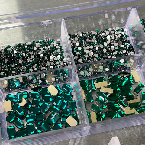 A clear organizer box from Dynamic Nail Supply securely holds an assortment of green gemstones. In the top section, it features small and round [Emerald Green] rhinestones reminiscent of emerald green nail supplies. The bottom compartment is filled with a mix of larger rectangular and square gems with gold backing, all neatly arranged in a 20-celled box that includes 14 big shapes and 6 round shapes, beautifully displayed on a metallic surface.