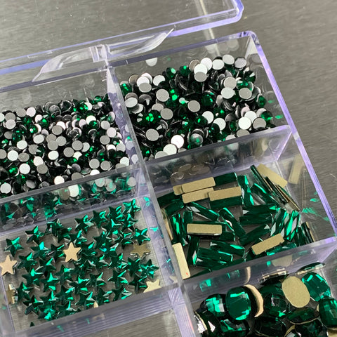 A clear plastic container from Dynamic Nail Supply is divided into four sections, each filled with a variety of green and silver craft embellishments. The sections hold round silver studs, emerald green rhinestones from the "Emerald Green Rhinestones Set," metallic star shapes, and elongated crystals, all neatly organized for crafting purposes.