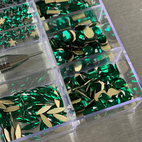 A close-up of the Emerald Green Rhinestones Set from Dynamic Nail Supply reveals various green and gold nail art decorations, meticulously organized in a transparent plastic case with 20 sections. The box features both star-shaped and faceted gems among its 14 big shapes and 6 round shapes. A metal tool with a fine tip is visible on the left against a metallic surface, making it an ideal choice for nail supplies enthusiasts.
