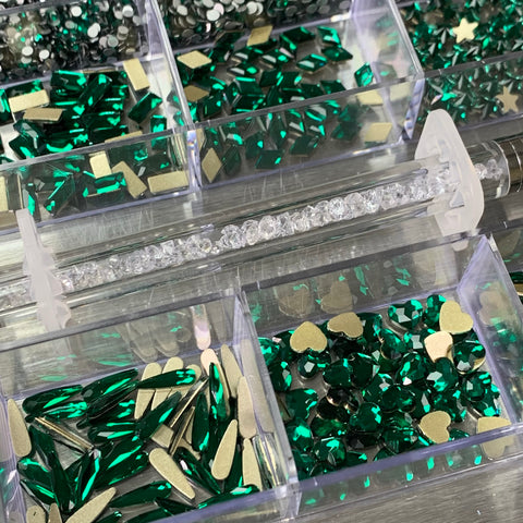 This Dynamic Nail Supply [Emerald Green] Rhinestones set includes a 20-cell box filled with various nail art decorations, offering a selection of green gemstones, emerald green rhinestones, gold hearts, stars, and teardrops. It also comes with a tube of clear rhinestones for boundless creativity.