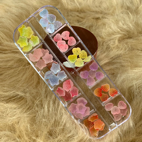 A rectangular container from Dynamic Nail Supply holds 48 pieces of star and heart-shaped gummy sugar charms for nail art design. They come in various vibrant colors, including shades of blue, pink, yellow, red, orange, and purple, which shimmer beautifully on a soft, fuzzy light brown surface.