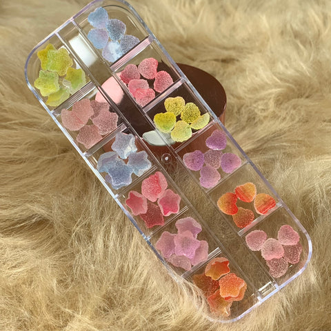 Dynamic Nail Supply's 48-piece set of star and heart-shaped gummy sugar charms is crafted for nail art design enthusiasts. These pastel-colored transparent plastic containers hold pink, yellow, purple, and orange charms neatly on a soft brown textured surface.