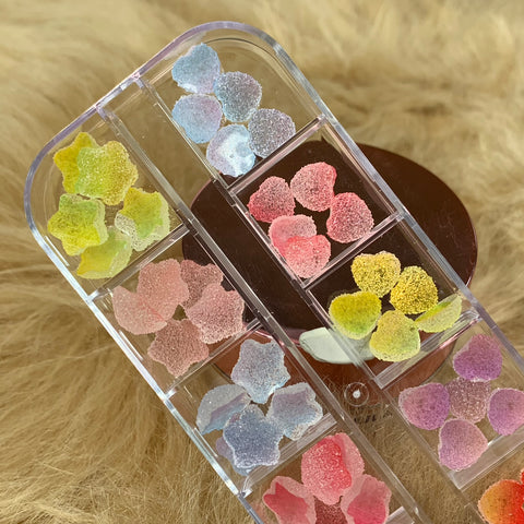 A clear organizer with several sections contains vibrant, Star and Heart shape Gummy sugar Charms perfect for nail art design. The pastel shades of yellow, pink, blue, and purple stand out beautifully against the organizer from Dynamic Nail Supply as it sits on a textured beige surface.