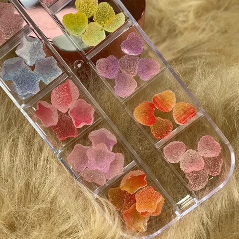 A close-up of a clear container featuring 48 vibrant gummy sugar charms in star and heart shapes, ideal for nail art design under the Dynamic Nail Supply brand. These colorful charms include shades like pink, purple, orange, yellow, and green. The container is set against a textured beige surface resembling fur or faux fur, creating a soft background.
