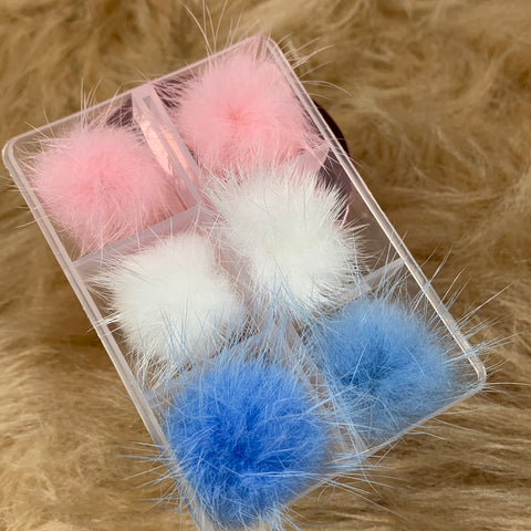 This clear plastic box from Dynamic Nail Supply contains three pairs of pom-poms for nail art design, featuring the Pompom version in soft, wispy textures with two in baby pink, two in white, and two in baby blue. They are set against a fuzzy beige surface, making them perfect for adding charm to your nail creations.