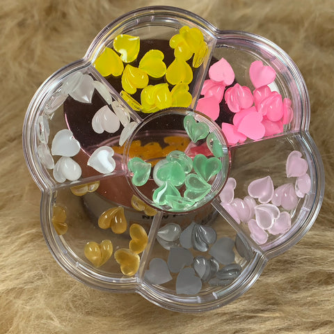 A clear plastic container in the shape of a flower features seven compartments, each filled with colorful resin heart-shaped charms in shades of yellow, pink, white, green, pale blue, gray, and gold. Ideal for crafting Valentines Nails designs or adding a touch of style to your appearance from Dynamic Nail Supply.