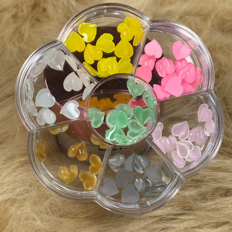 A flower-shaped container houses heart-shaped beads in an array of vibrant colors—yellow, pink, light pink, brown, white, and green. This delightful arrangement is set against a fluffy beige fur surface, making it ideal for crafting Valentines Nails or combining with Dynamic Nail Supply's Colorful Resin Heart-Shaped Charms for Valentine's Nails Art Design.