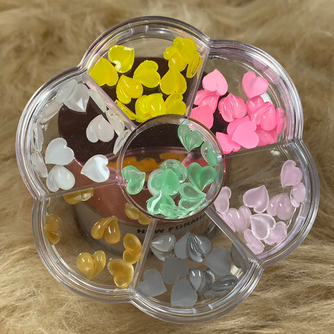 The Dynamic Nail Supply presents a flower-shaped container featuring six compartments filled with vibrant, heart-shaped beads in yellow, pink, and lavender hues. These colorful resin heart-shaped charms are perfect for crafting stunning Valentine's nail designs against a textured, light brown fur-like backdrop.