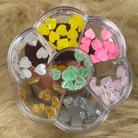 The flower-shaped plastic container from Dynamic Nail Supply, featuring six compartments, is filled with Colorful Resin Heart-shaped charms and heart-shaped beads in a palette of yellow, pink, white, brown, green, and gray. Ideal for crafting or designing unique Valentine's nails, the container elegantly rests on a textured brown surface.